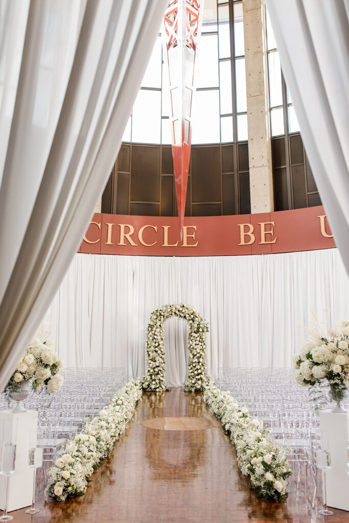Curtained entryway of ceremony space with view of floral arbor and floral lined aisle.