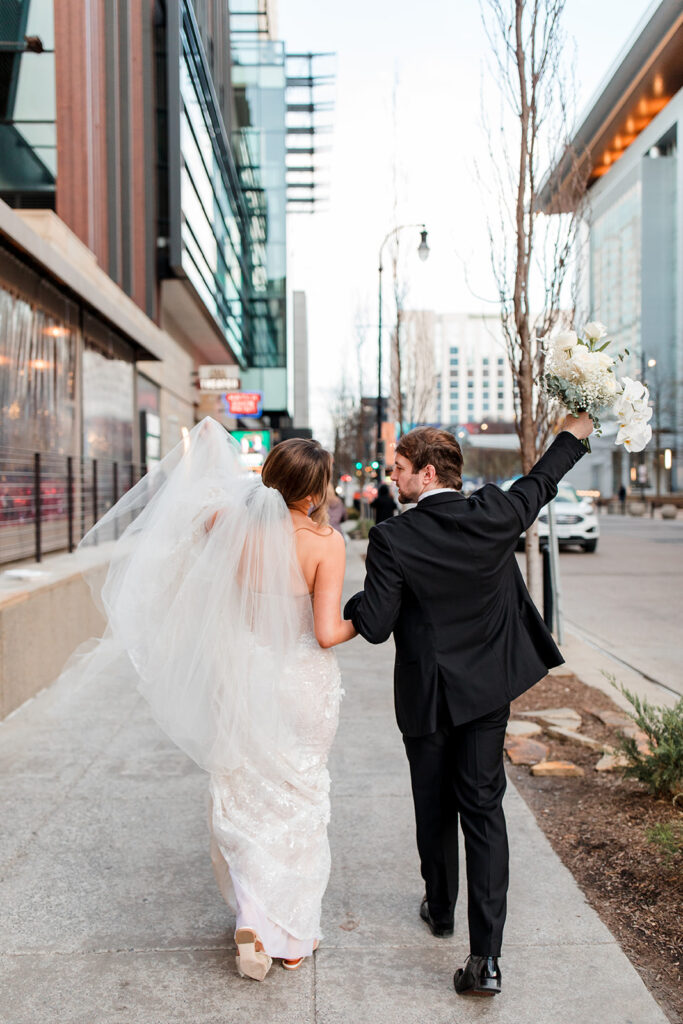 Bride and Groom walking down the street. Groom holding bouquet in the air. 