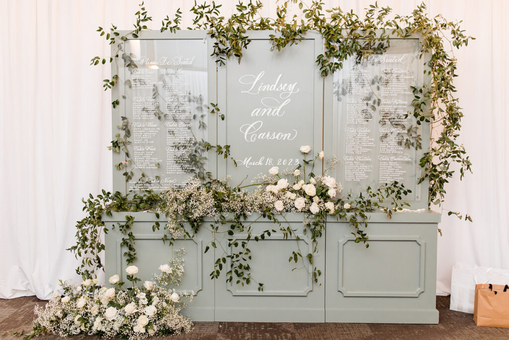 Florals cascading down a soft gray, wooden seating chart with white ink on acrylic signage