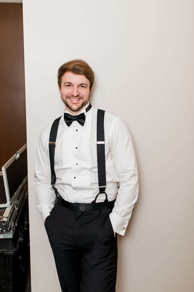 Groom smiling with hands in pockets, leaning against a wall.