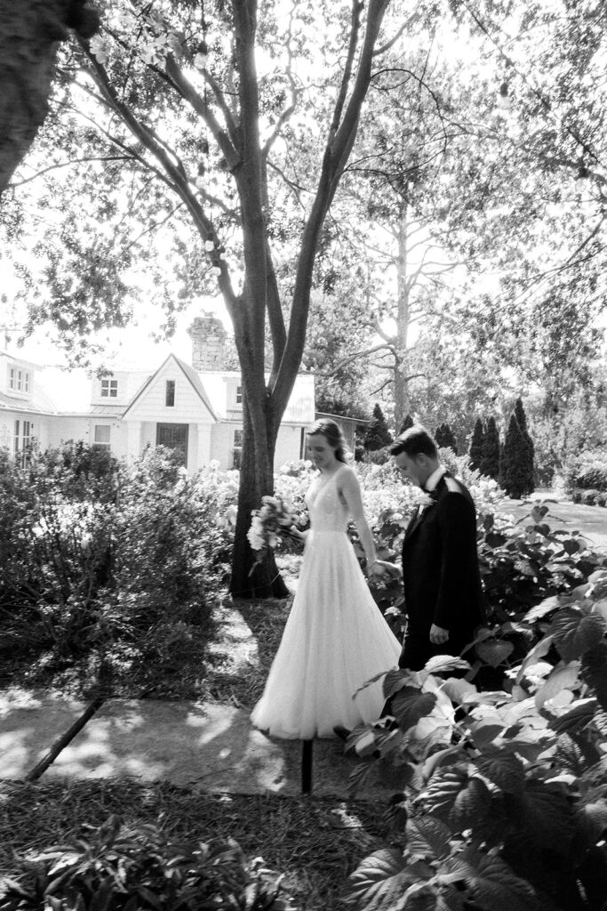 Bride and groom walking along on path among plants and flowers with cottage in the background.