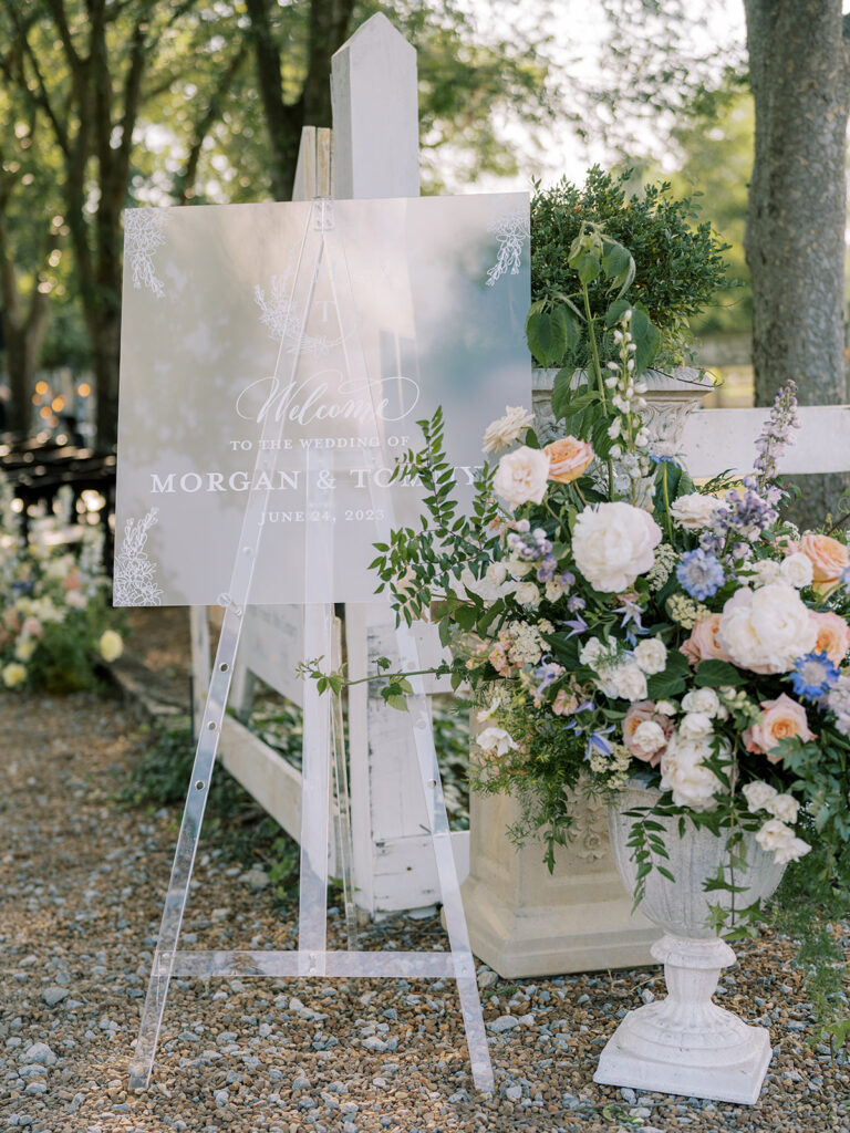 Outdoor frosted acrylic wedding welcome sign on easel with flower arrangement in front.