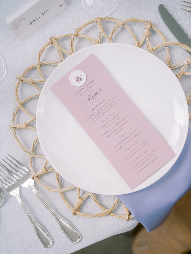 Wax seal atop lavender reception menu placed over white plate on wicker charger.