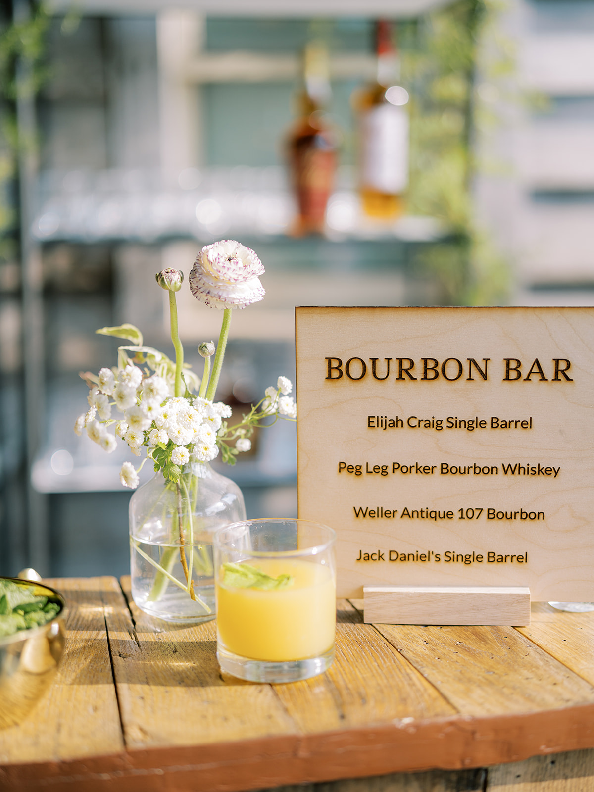 Engraved wooden bar sign on top of bar with prepared drink and small flower arrangment.