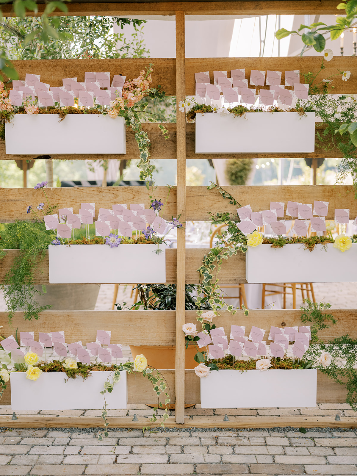 Escort card display with escort cards placed inside wooden boxes on wooden shelving decorated in florals and greenery. Dreamy Garden-Inspired Summer Wedding details.