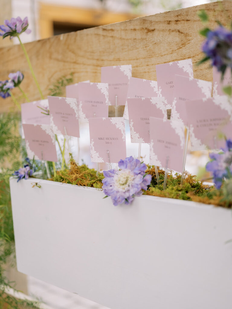 Escort card display with escort cards placed inside wooden boxes on wooden shelving decorated in florals and greenery.