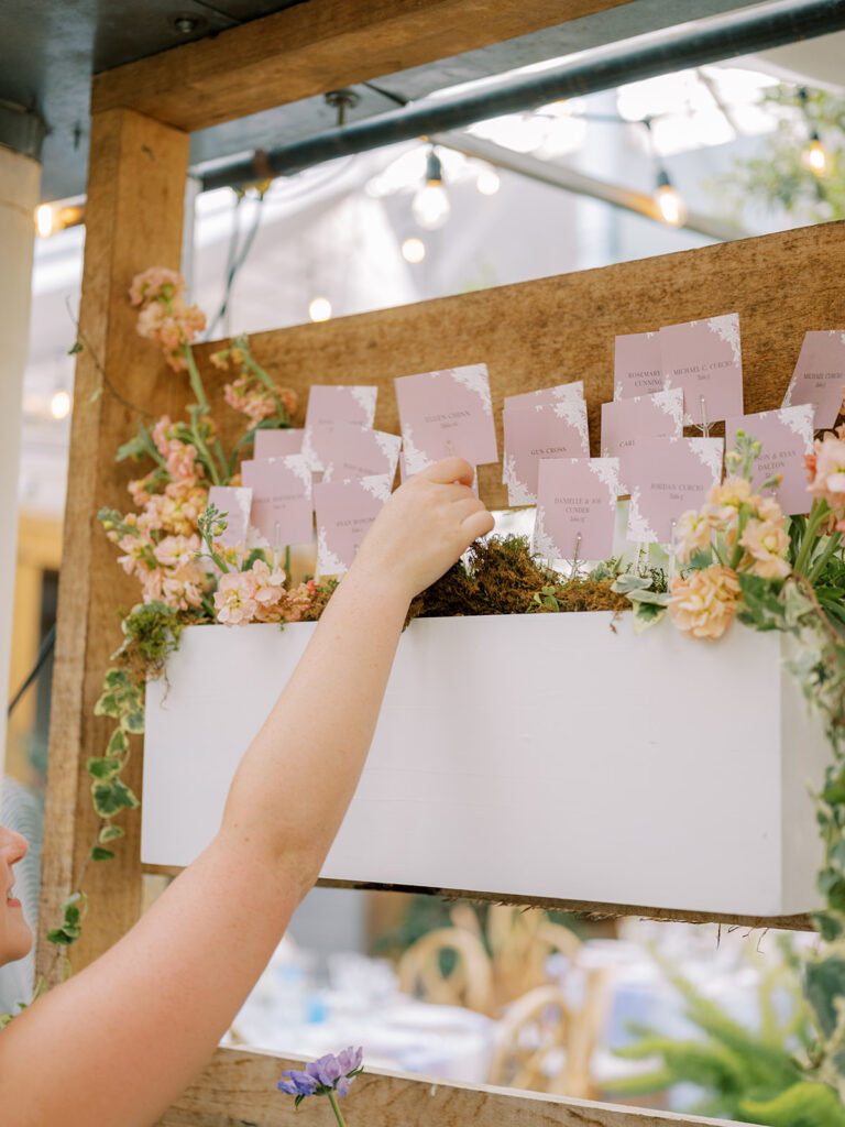 Escort card display with escort cards placed inside wooden boxes on wooden shelving decorated in florals and greenery. Person's arm reaching to pull out escort card.