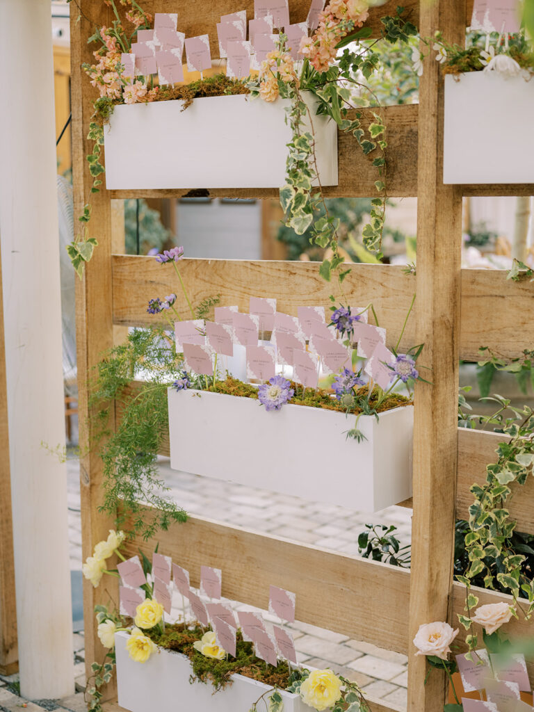 Escort card display with escort cards placed inside wooden boxes on wooden shelving decorated in florals and greenery.