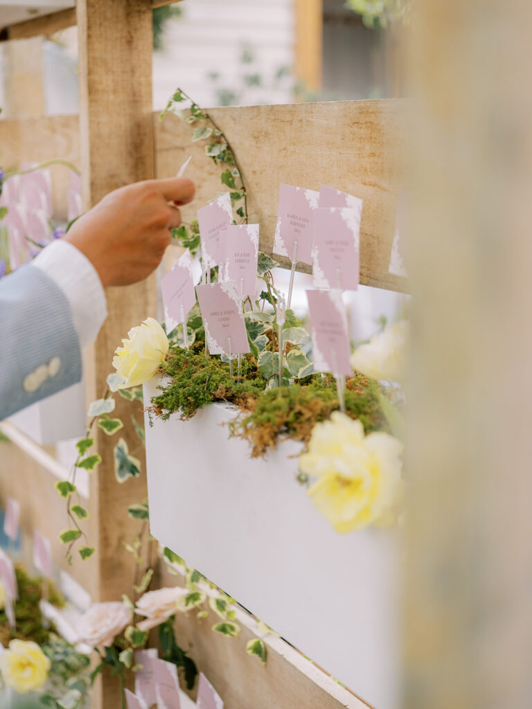 Escort card display with escort cards placed inside wooden boxes on wooden shelving decorated in florals and greenery. Person's hand reaching to pull out escort card.