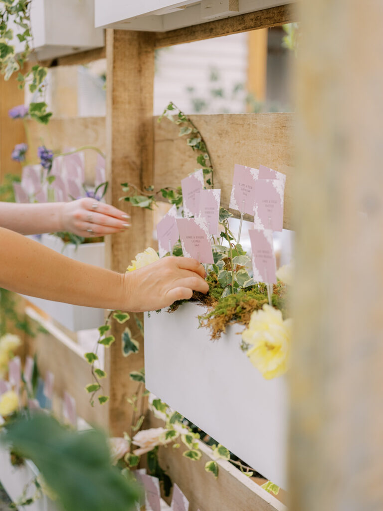 Escort card display with escort cards placed inside wooden boxes on wooden shelving decorated in florals and greenery. People's hands reaching to pull out escort card.