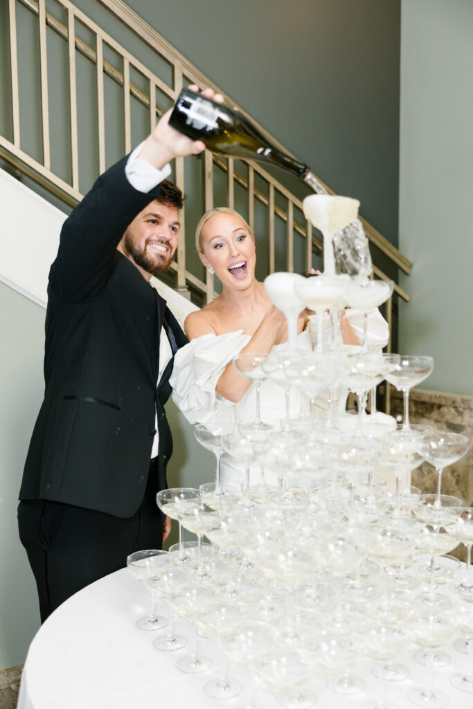 Bride and groom standing cheerfully while groom pours champagne of tower of champagne glasses. 