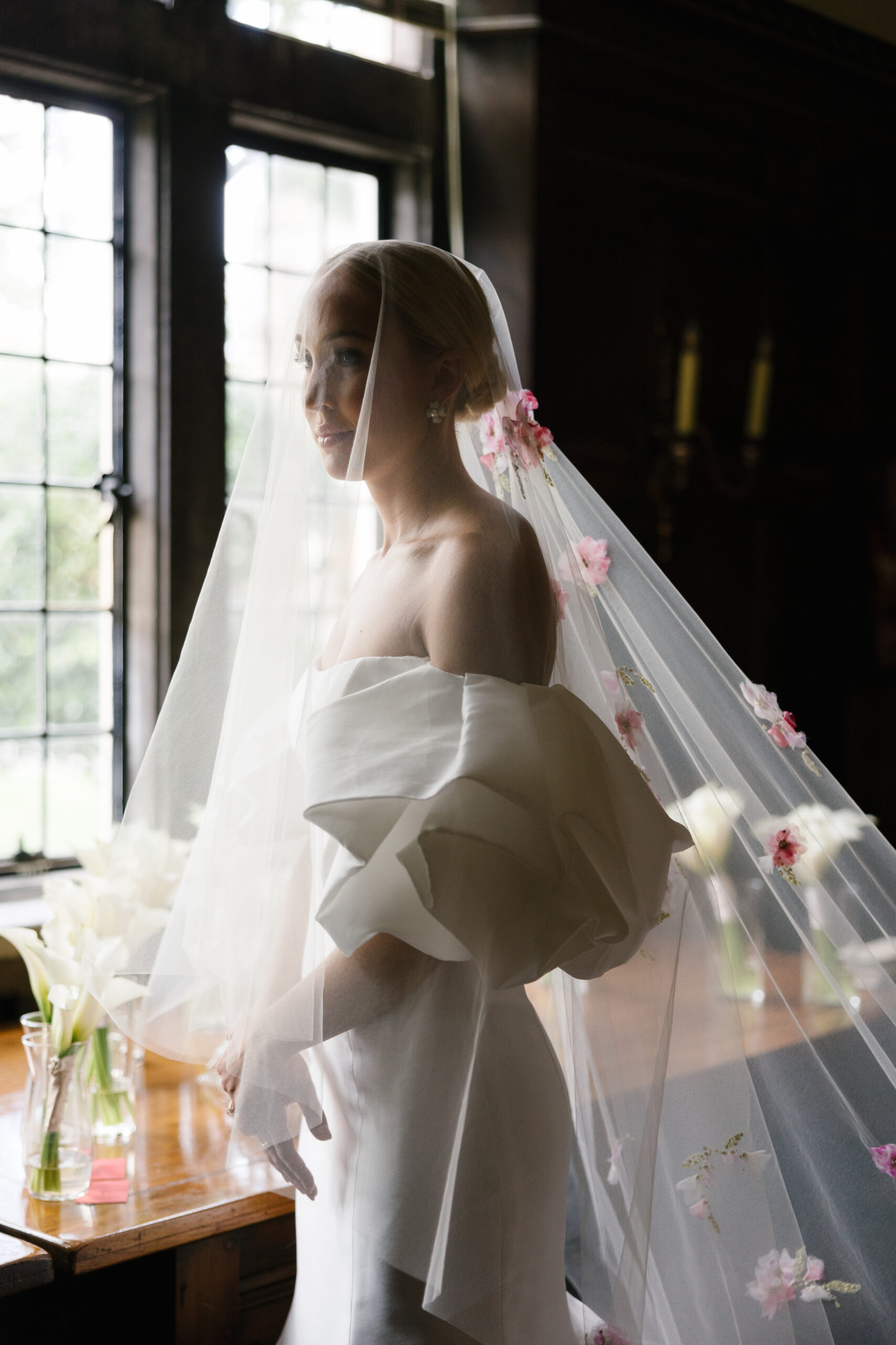 Bride dressed in gown and veil looking out of window.