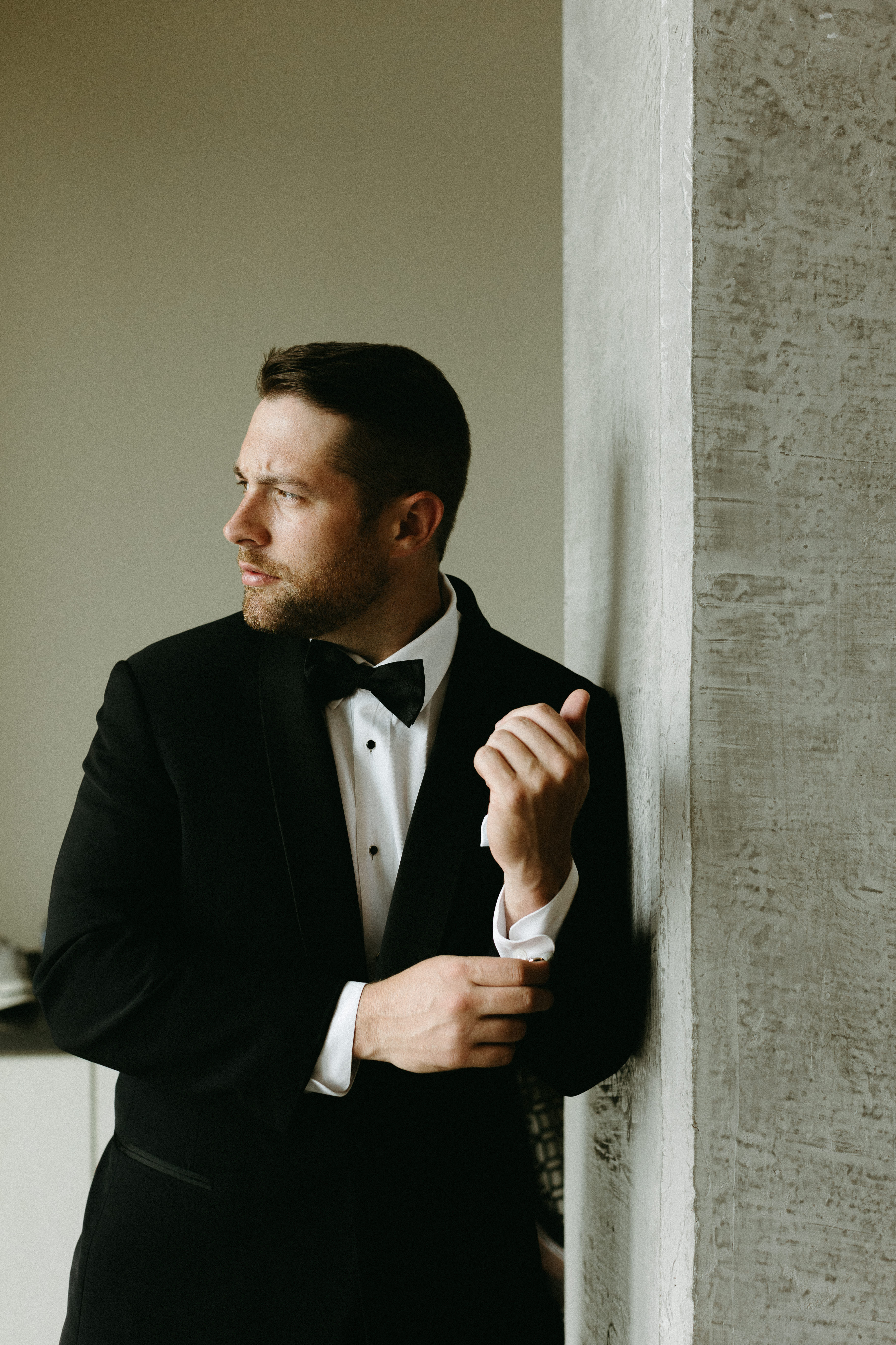 Groom in a black tux leaning against a wall looking off to the side while adjusting his cuff. 