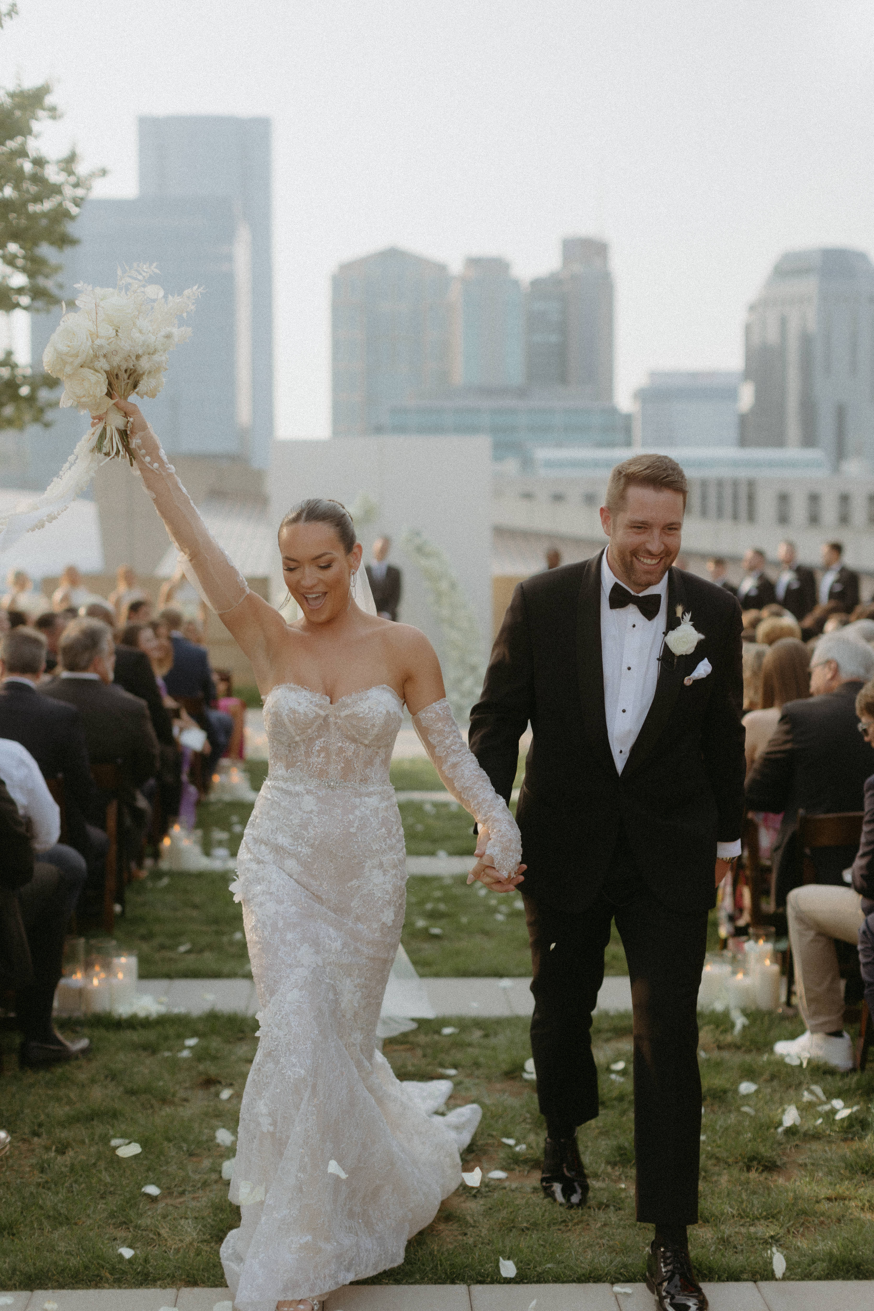 Bride and Groom walking down the outdoor aisle with Nashville skyline in the background. Bride is hoisting up bouquet of flowers. 