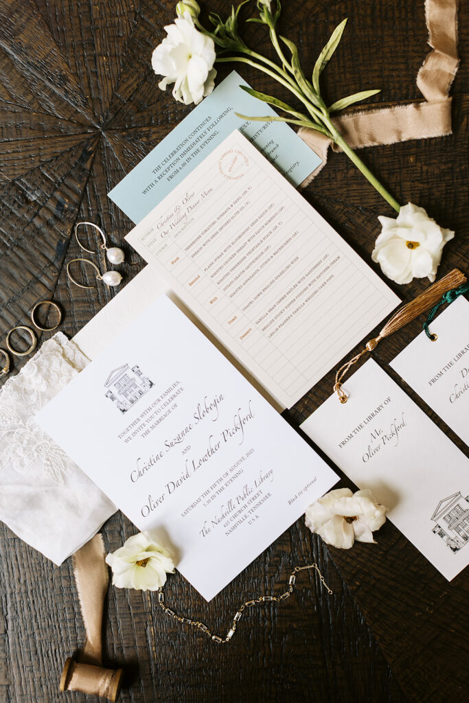 Wedding invitation suite showcasing custom sketch and styled with jewelry and florals. Sitting on wooden surface.