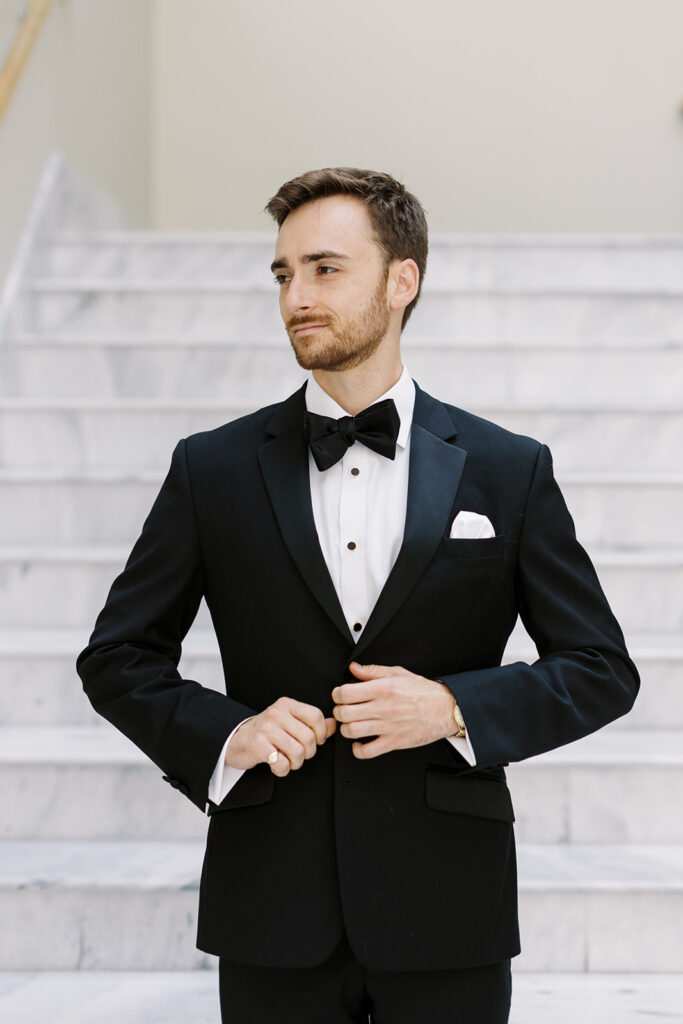Groom on stairs buttoning his tux looking to the side.