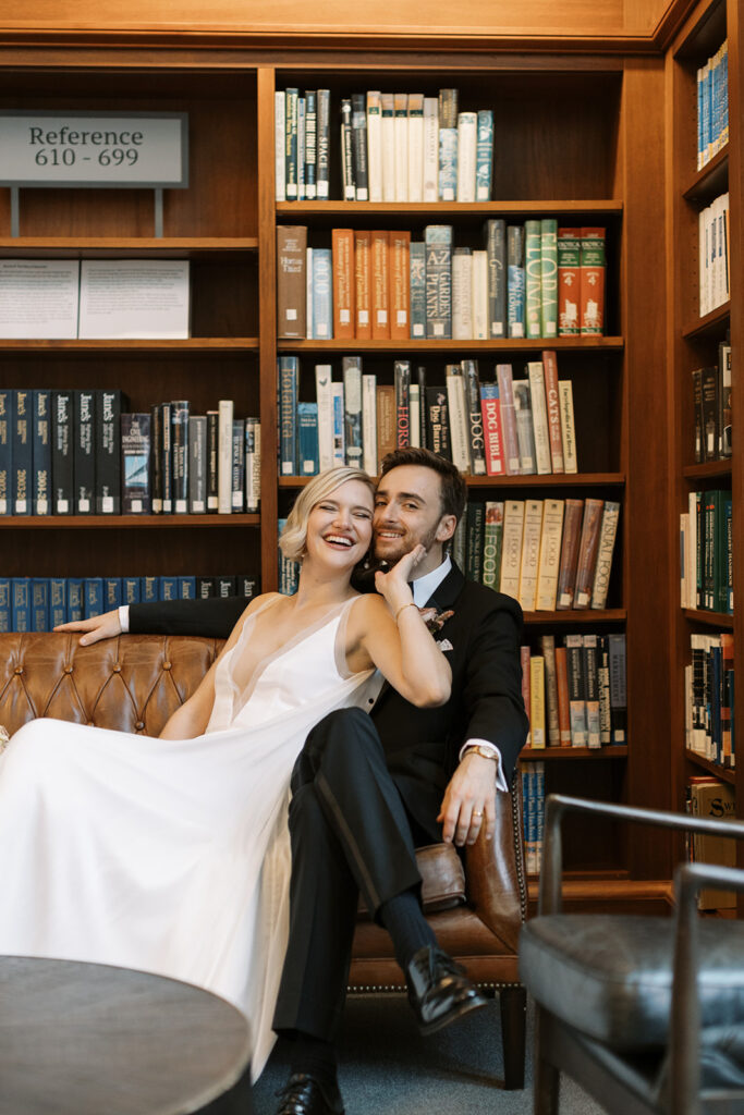 Bride and Groom laughing together while seated on a sofa in front of book shelves. 
