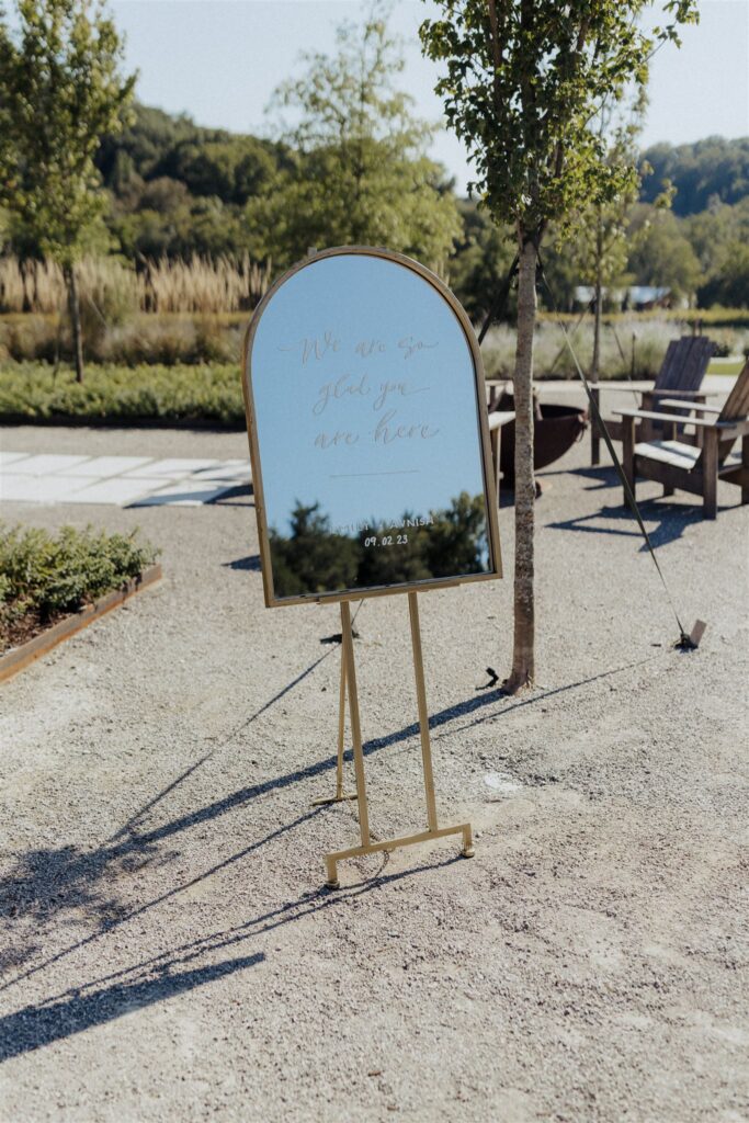 Arched, mirrored wedding welcome sign standing amidst outdoor venue.