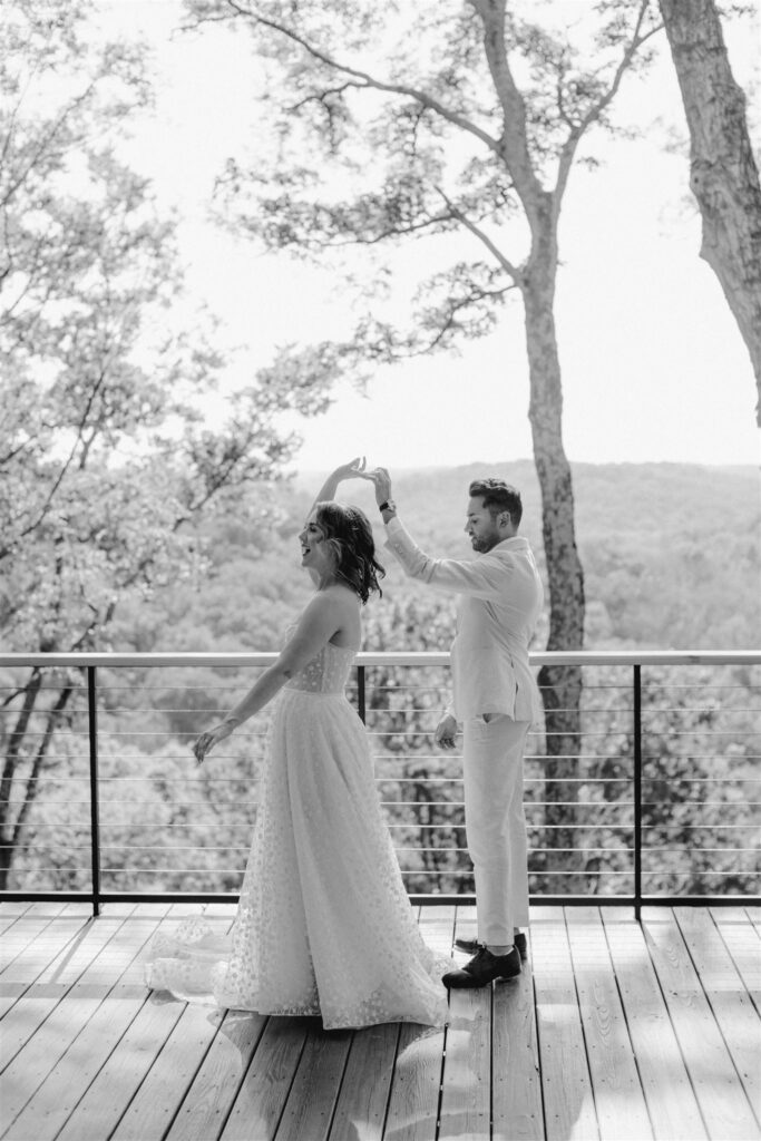 Groom spinning bride around on outdoor balcony with treeline in the background.