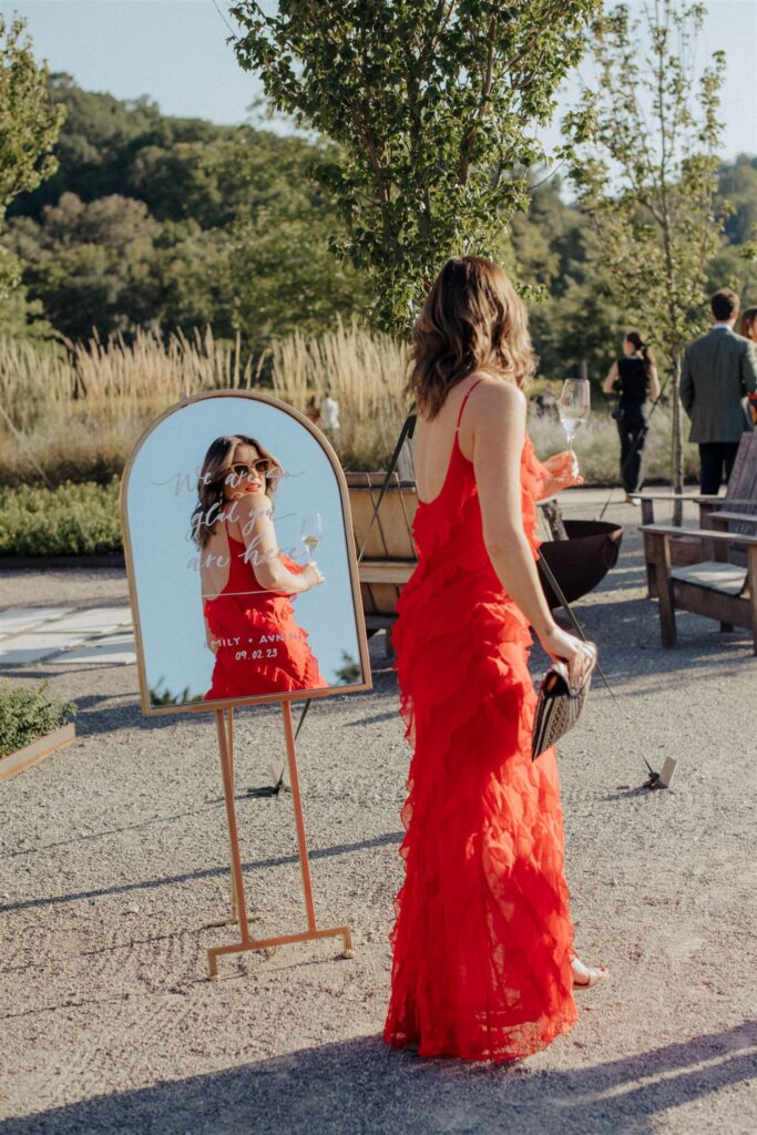 Woman in red dress looking at her reflection in outdoor, mirrored wedding welcome sign.