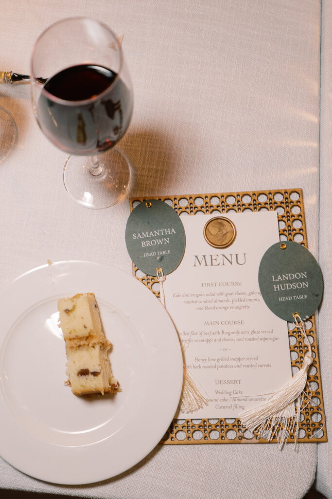Wedding reception menu and escort card sits beside plate of food and glass of wine.