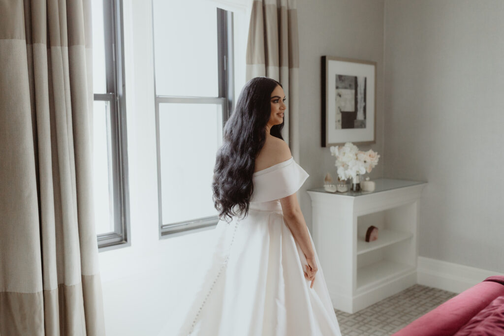 Bride in gown, long dark hair, standing in front of window