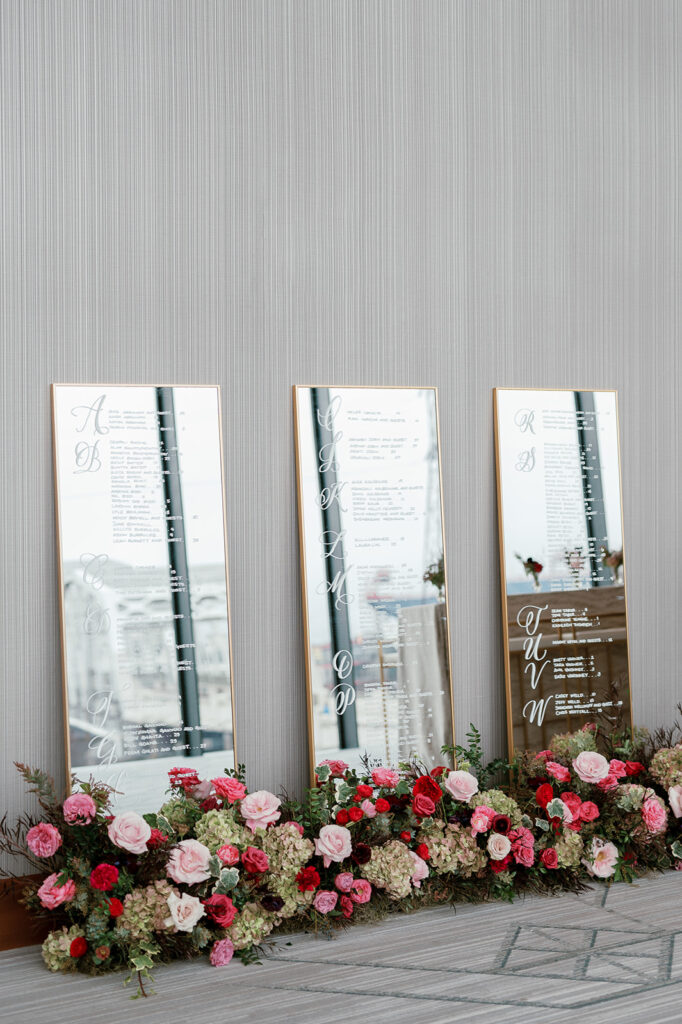 Row of mirrored seating charts standing against a wall. Surrounded by florals at their base.