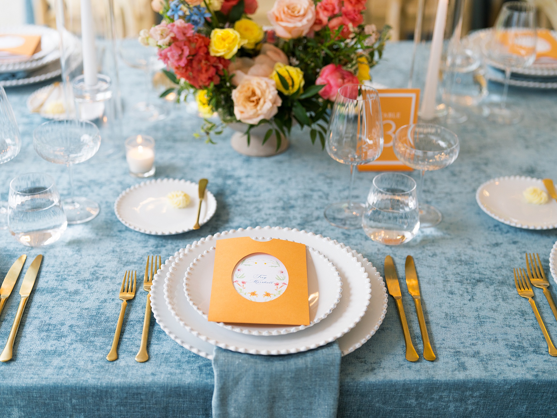 menus tucked into a custom die-cut orange pocket, featuring floral artwork. The menus and orange acrylic table numbers popped against the blue linens and tied into the colorful spring floral centerpieces. 